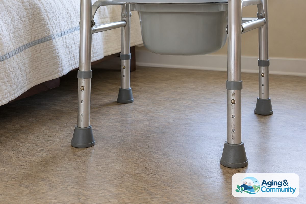 The lower legs of a commode stand with four rubber feet flat on a level vinyl floor beside a bed.